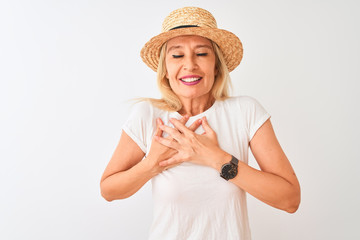 Middle age woman wearing casual t-shirt and hat standing over isolated white background smiling with hands on chest with closed eyes and grateful gesture on face. Health concept.