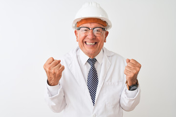 Senior grey-haired engineer man wearing coat and helmet over isolated white background celebrating...