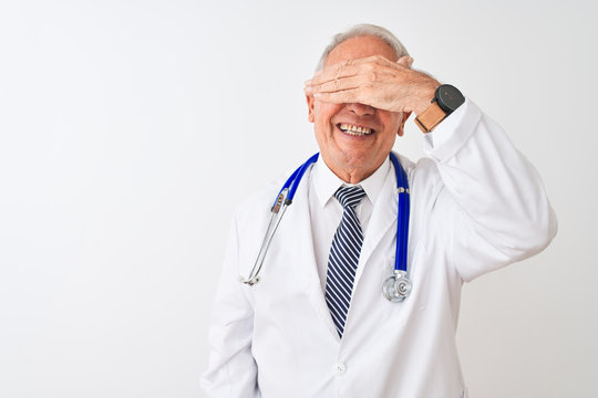 Senior grey-haired doctor man wearing stethoscope standing over isolated white background smiling and laughing with hand on face covering eyes for surprise. Blind concept.