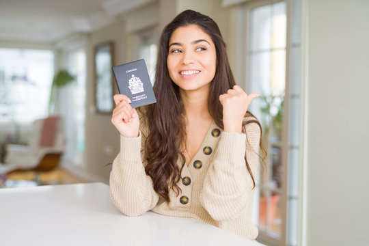 Young woman holding passport of Canada pointing and showing with thumb up to the side with happy face smiling