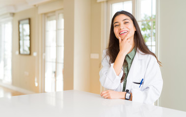 Young woman wearing medical coat at the clinic as therapist or doctor looking confident at the camera with smile with crossed arms and hand raised on chin. Thinking positive.