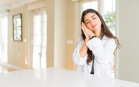 Young woman wearing medical coat at the clinic as therapist or doctor sleeping tired dreaming and posing with hands together while smiling with closed eyes.
