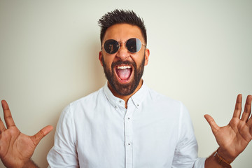 Handsome indian buinessman wearing shirt and sunglasses over isolated white background celebrating mad and crazy for success with arms raised and closed eyes screaming excited. Winner concept