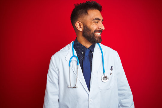 Young indian doctor man wearing stethoscope standing over isolated red background looking away to side with smile on face, natural expression. Laughing confident.
