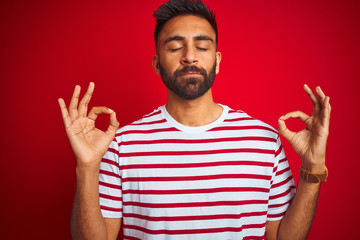 Young indian man wearing striped t-shirt standing over isolated red background relax and smiling with eyes closed doing meditation gesture with fingers. Yoga concept.