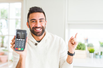 Handsome hispanic man holding point of sale terminal dataphone very happy pointing with hand and finger to the side