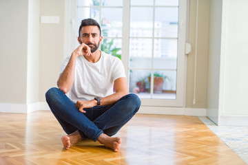 Handsome hispanic man wearing casual t-shirt sitting on the floor at home with hand on chin thinking about question, pensive expression. Smiling with thoughtful face. Doubt concept.