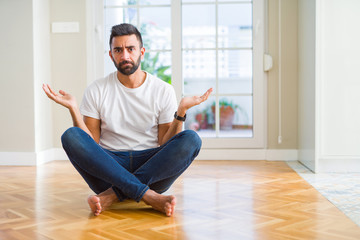 Handsome hispanic man wearing casual t-shirt sitting on the floor at home clueless and confused expression with arms and hands raised. Doubt concept.