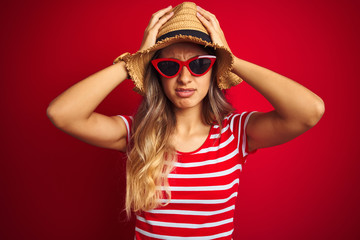 Young beautiful woman wearing sunglasses and summer hat over red isolated background suffering from headache desperate and stressed because pain and migraine. Hands on head.