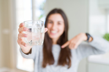 Beautiful young woman drinking a fresh glass of water with surprise face pointing finger to himself