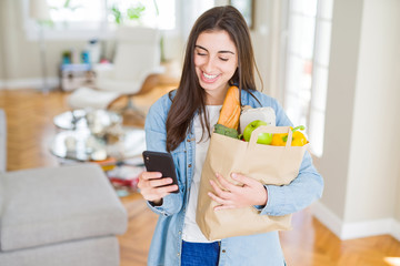 Young woman holding a paper bag full of fresh groceries and using smartphone app for supermarket...