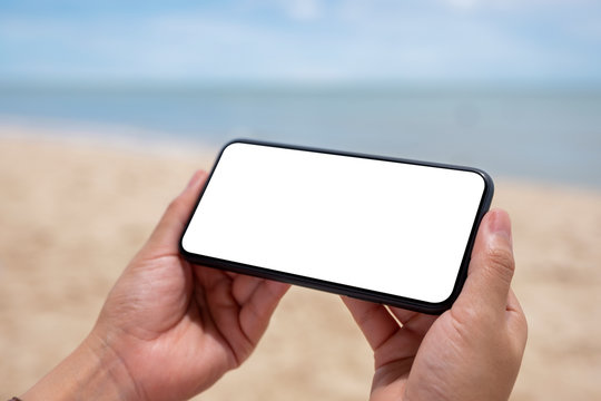 Mockup image of hands holding black mobile phone with blank desktop screen by the sea