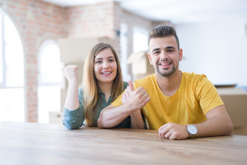 Young couple sitting on the table movinto to new home with carboard boxes behind them smiling with happy face looking and pointing to the side with thumb up.