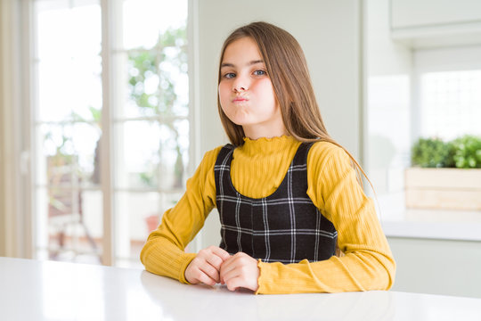 Young beautiful blonde kid girl wearing casual yellow sweater at home puffing cheeks with funny face. Mouth inflated with air, crazy expression.
