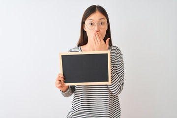 Young chinese woman wearing glasses holding blackboard over isolated white background cover mouth with hand shocked with shame for mistake, expression of fear, scared in silence, secret concept