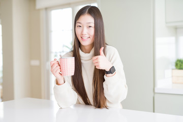 Beautiful Asian woman drinking a cup of coffee happy with big smile doing ok sign, thumb up with fingers, excellent sign