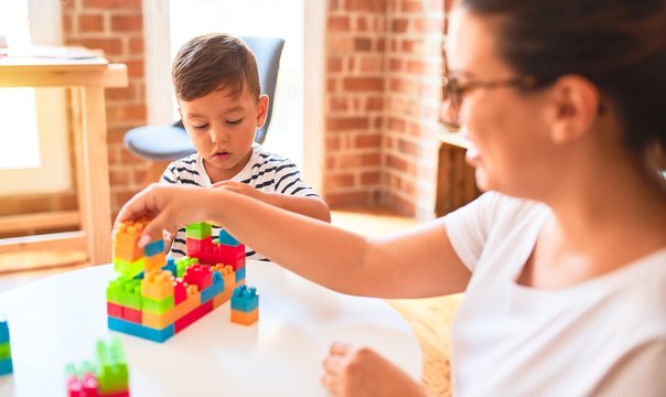 Beautiful teacher and toddler boy playing with construction blocks bulding tower at kindergarten