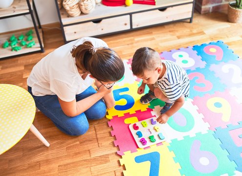 Beautiful teacher and toddler boy sitting on puzzle playing with numbers at kindergarten