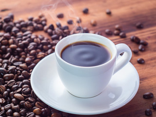 Smoking aromatic espresso in white coffee cup, close up view. Roasted coffee beans pilled on brown table. Texture of wooden desk. Selective soft focus. Blurred background