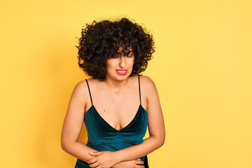 Young arab woman with curly hair wearing elegant dress over isolated yellow background with hand on stomach because nausea, painful disease feeling unwell. Ache concept.