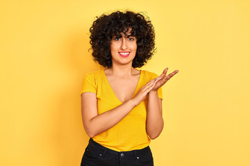 Young arab woman with curly hair wearing t-shirt standing over isolated yellow background clapping...