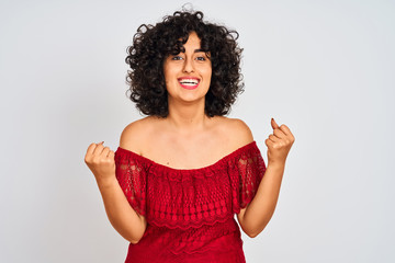 Young arab woman with curly hair wearing red dress standing over isolated white background celebrating surprised and amazed for success with arms raised and open eyes. Winner concept.