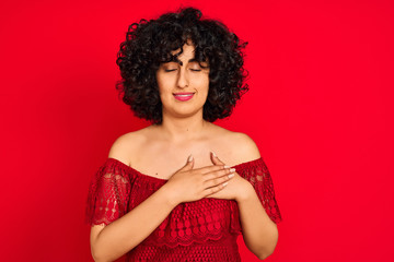 Young arab woman with curly hair wearing casual dress over isolated red background smiling with hands on chest with closed eyes and grateful gesture on face. Health concept.