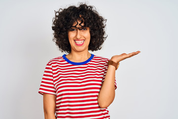 Young arab woman with curly hair wearing striped t-shirt over isolated white background smiling cheerful presenting and pointing with palm of hand looking at the camera.