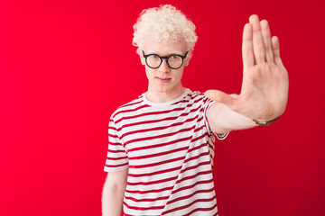 Young albino blond man wearing striped t-shirt and glasses over isolated red background doing stop sing with palm of the hand. Warning expression with negative and serious gesture on the face.