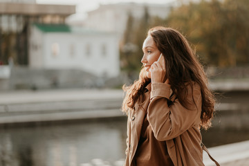 Charming smiling brunette woman with long curly hair dressed casual coat with mobile phone in hands at city street