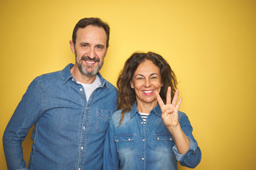 Naklejka premium Beautiful middle age couple together standing over isolated yellow background showing and pointing up with fingers number four while smiling confident and happy.