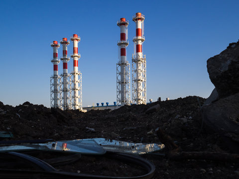 Red-and-white boiler room chimneys against a blue sky. Mountain of stones and debris and pipes boiler station