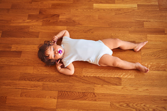 Beautiful toddler child girl wearing white bodysuit lying down on the floor using pacifier