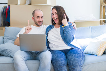 Young couple sitting on the sofa arround cardboard boxes moving to a new house using laptop surprised with an idea or question pointing finger with happy face, number one