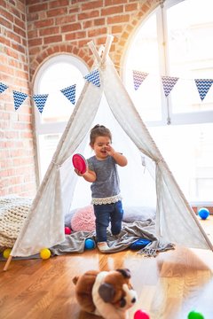 Beautiful toddler standing inside tipi holding plastic dishes smiling at kindergarten