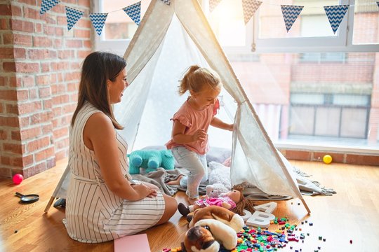 Beautiful teacher and blond toddler girl playing with dolls inside tipi at kindergarten