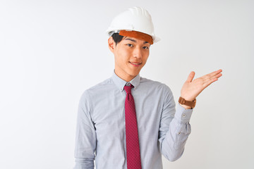 Chinese architect man wearing tie and helmet standing over isolated white background smiling cheerful presenting and pointing with palm of hand looking at the camera.