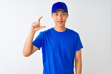 Chinese deliveryman wearing blue t-shirt and cap standing over isolated white background smiling and confident gesturing with hand doing small size sign with fingers looking and the camera