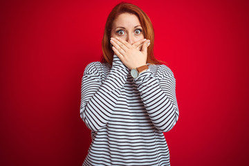 Young redhead woman wearing strapes navy shirt standing over red isolated background shocked covering mouth with hands for mistake. Secret concept.