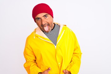Middle age man wearing rain coat and woolen hat standing over isolated white background sticking tongue out happy with funny expression. Emotion concept.