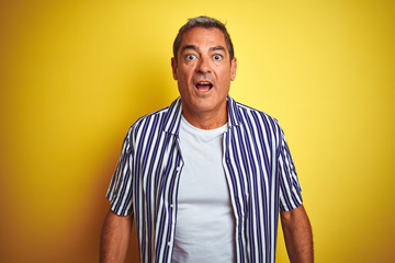 Handsome middle age man wearing striped shirt standing over isolated yellow background afraid and shocked with surprise expression, fear and excited face.