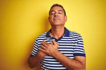 Handsome middle age man wearing striped polo standing over isolated yellow background smiling with hands on chest with closed eyes and grateful gesture on face. Health concept.