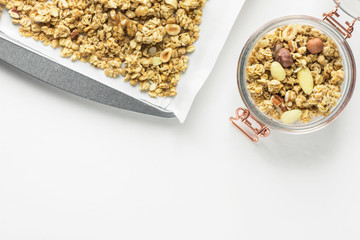 Homemade Granola Muesli with oats dried fruits coconut flakes hazelnuts almonds walnuts mixture on baking tray and in glass jar on white table. Healthy wholesome food fiber energy source