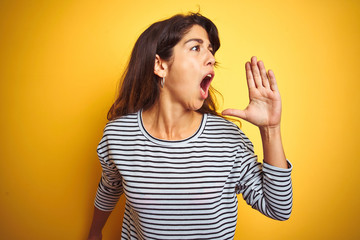 Young beautiful woman wearing stripes t-shirt standing over yelllow isolated background shouting and screaming loud to side with hand on mouth. Communication concept.