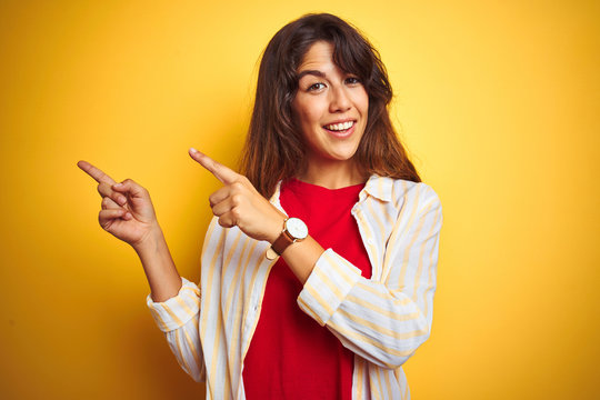 Young beautiful woman wearing red t-shirt and stripes shirt over yellow isolated background smiling and looking at the camera pointing with two hands and fingers to the side.