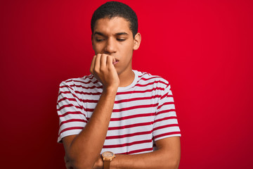 Young handsome arab man wearing striped t-shirt over isolated red background looking stressed and nervous with hands on mouth biting nails. Anxiety problem.