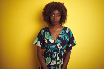 Young african afro woman wearing summer floral dress over isolated yellow background with serious expression on face. Simple and natural looking at the camera.