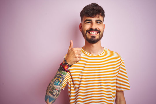 Young man with tattoo wearing yellow striped t-shirt standing over isolated pink background doing happy thumbs up gesture with hand. Approving expression looking at the camera with showing success.