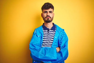 Young handsome man wearing rain coat standing over isolated yellow background skeptic and nervous, disapproving expression on face with crossed arms. Negative person.