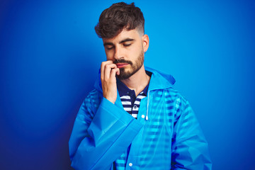 Young handsome man wearing rain coat standing over isolated blue background thinking looking tired and bored with depression problems with crossed arms.
